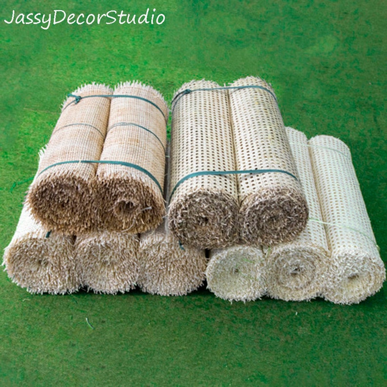 May include: Several rolls of woven rattan material in varying shades of beige and brown are stacked on a green surface. The rolls are secured with green twine. The text "JassyDecorStudio" is in the upper left corner.