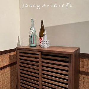 May include: A wooden cabinet with slatted doors, featuring two bottles and a decorative ceramic object on top. The cabinet has a brown wood finish and the bottles are clear glass with labels. The ceramic object is white with a blue eye and a spiky design.
