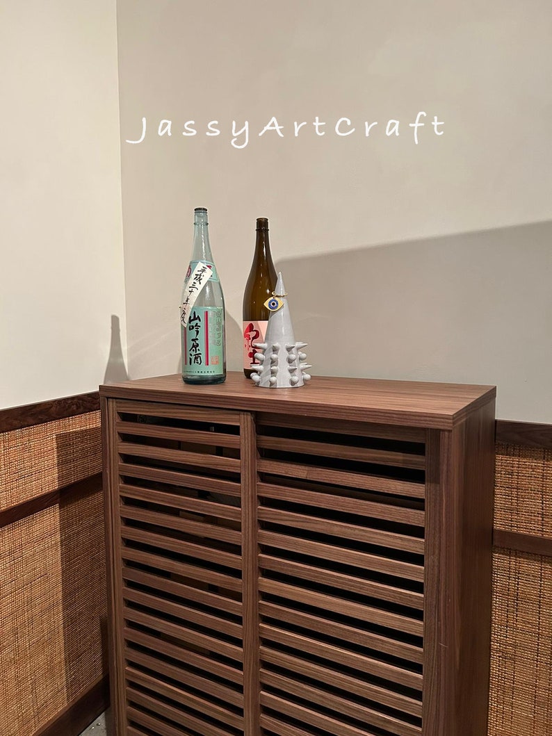 May include: A wooden cabinet with slatted doors, featuring two bottles and a decorative ceramic object on top. The cabinet has a brown wood finish and the bottles are clear glass with labels. The ceramic object is white with a blue eye and a spiky design.