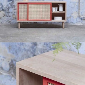 May include: A light wood cabinet with a red-framed, woven cane sliding door. The cabinet has a shelf and is topped with a small, beige lamp. The cabinet is on short legs and has a minimalist design. The background is a textured wall.