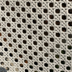 May include: Close-up of a white woven cane pattern. The design features a repeating geometric grid of interwoven strips, creating a textured surface. The background shows glimpses of a darker, natural material.