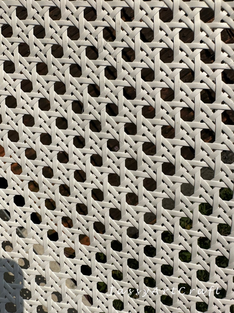 May include: Close-up of a white woven cane pattern. The design features a repeating geometric grid of interwoven strips, creating a textured surface. The background shows glimpses of a darker, natural material.