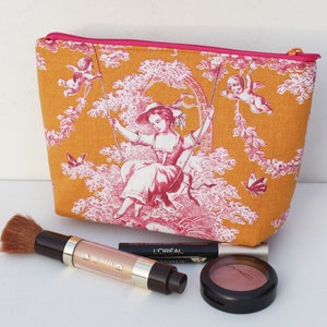 Könnte beinhalten: Eine Kosmetiktasche aus gelbem Toile mit einem rosafarbenen Blumenmuster und einem rosafarbenen Reißverschluss. Die Tasche zeigt eine Szene mit einer Frau auf einer Schaukel mit zwei Cherubim.