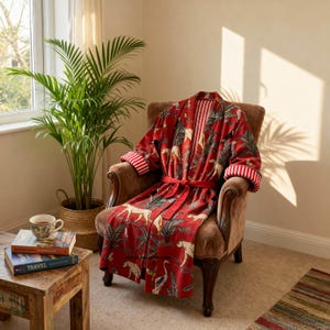 May include: A red robe featuring a repeating pattern of cheetahs and palm trees, with a matching tie belt and white and red striped cuffs. The robe has a folded collar and long sleeves, showcasing a vibrant jungle-inspired print throughout.
