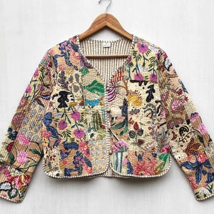 May include: A cream-coloured quilted jacket with a floral print. The jacket has a V-neck and long sleeves. The fabric is a patchwork of different floral patterns in shades of pink, blue, green, and yellow.