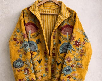 May include: A mustard yellow jacket featuring intricate floral embroidery in shades of red, blue, and green. The garment has long sleeves, a collared neckline, and an open front design. The interior lining displays a white and yellow striped pattern.