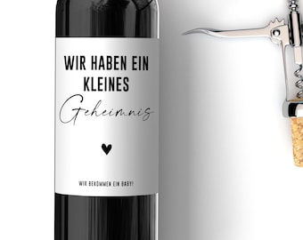 Può includere: Una bottiglia di vino nera con un'etichetta bianca che dice "Wir haben ein kleines Geheimnis" e "Wir bekommen ein Baby!" L'etichetta ha un piccolo cuore nero. Uno cavatappi argentato è sullo sfondo.