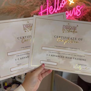 May include: Two certificates of completion for 1-1 advanced hair training. The certificates have gold lettering and a gold border. The certificates are held in front of a neon sign that says "Hello Girls" in pink.