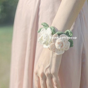 Op de afbeelding: Een gehaakt bloemenarmband met witte en roze bloemen en groene bladeren. De armband wordt om de pols gedragen.