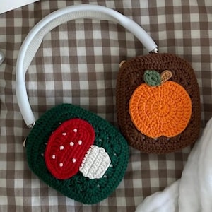 May include: Crocheted headphone covers, one with a red mushroom design and the other with an orange pumpkin. The covers are green and brown, respectively, and are connected by a white headband. The background includes a book and glasses.