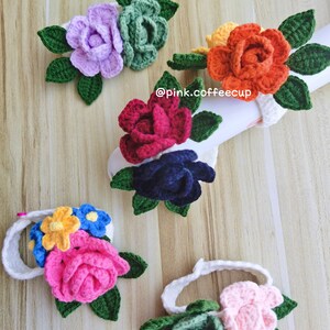 Op de afbeelding: Vijf gehaakte bloemenhaarbandjes in verschillende kleuren, waaronder roze, rood, paars, oranje en wit. Elk haarbandje is versierd met een gehaakte bloem met groene bladeren.