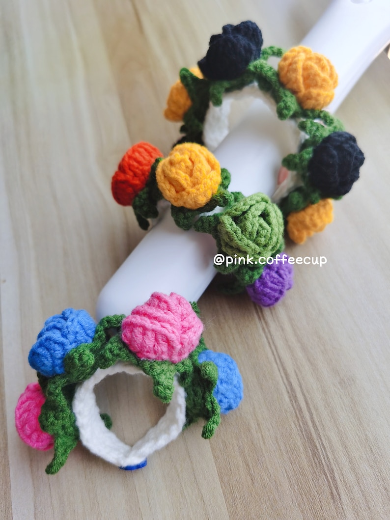 Op de afbeelding: Twee gebreide bloemen haarbandjes. De eerste haarband heeft zwarte, gele, oranje, groene en paarse bloemen. De tweede haarband heeft roze, blauwe en groene bloemen. De haarbandjes liggen op een witte achtergrond met de tekst "@pink.coffeecup".
