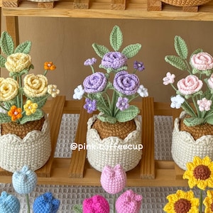 Op de afbeelding: Drie gehaakte bloemstukken in potten, elk met een ander kleurenschema: geel, paars en roze. De potten zijn allemaal wit en hebben een gegolfde rand. De bloemen zijn op een houten plank gerangschikt.