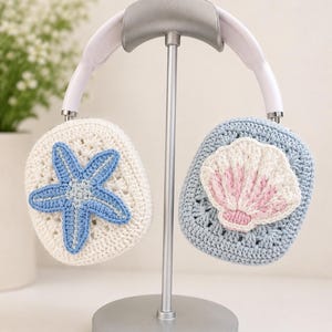 May include: White and light blue crochet headphone covers featuring a textured starfish applique on the left side and a seashell applique with pink accents on the right side, displayed on a silver metal headphone stand.