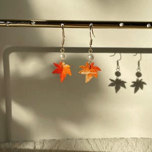 May include: Dangle earrings with orange and yellow maple leaf charms and a pearl bead. The earrings hang from gold-coloured hooks. The earrings are displayed on a gold-coloured bar.