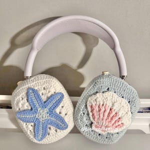 Może przedstawiać: Para słuchawek wykonana z białej i niebieskiej włóczki z motywem gwiazdki morskiej i muszli. Pokrowiec jest wykonany z miękkiej przędzy i idealnie nadaje się do ochrony słuchawek przed zarysowaniami i brudem.