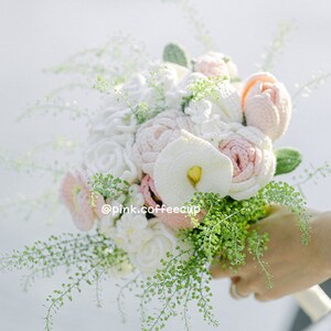 Op de afbeelding: Een gehaakt bruidsboeket met witte en roze bloemen en groen blad. Het boeket wordt vastgehouden door een hand en de tekst "@pink.coffeecup" is zichtbaar.