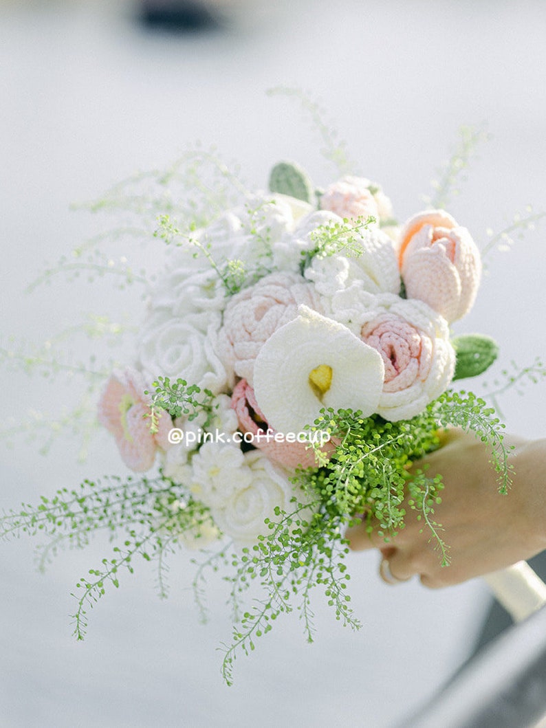 Op de afbeelding: Een gehaakt bruidsboeket met witte en roze bloemen en groen blad. Het boeket wordt vastgehouden door een hand en de tekst "@pink.coffeecup" is zichtbaar.