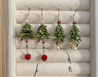 May include: Four pairs of Christmas tree earrings. The earrings are green with red and gold accents, and feature gold-colored hooks. Some earrings have red, faceted bead drops, while others have gold leaf and pearl accents. The earrings are displayed in a jewelry box.