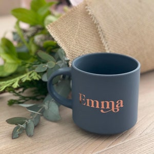May include: A dark grey ceramic mug with a rose gold inscription that reads "Emma".
