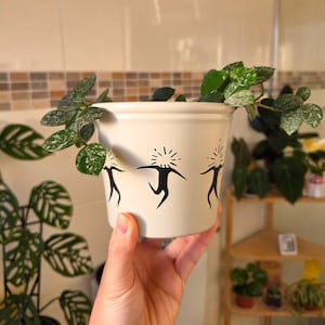 May include: A cream-coloured ceramic plant pot featuring a black graphic of stylized figures. The pot is filled with a green plant with speckled leaves. The pot is held in a person's hand.