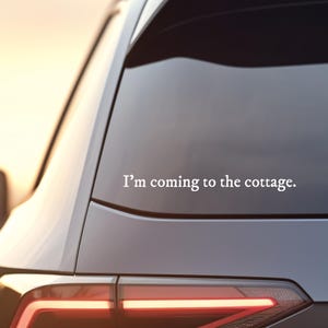May include: A white sticker on a car window displaying the text "I'm coming to the cottage." The car is dark grey with a red taillight. The background shows a blurred sunset.