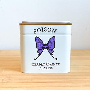 May include: A white rectangular tin container with a gold-coloured lid. The front features the word POISON in black text above a purple butterfly graphic with a skull centre. Below the butterfly, the text reads DEADLY AGAINST DEMONS in black.