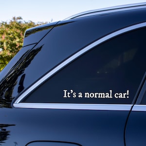 May include: A dark blue vehicle window featuring a white vinyl decal with the text It's a normal car! in a serif font. The sticker is applied to the rear side window of the car, which has silver trim along the window frame and a glossy finish.