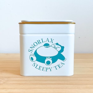 May include: A white rectangular metal tea tin with a gold-coloured lid. The front features a teal graphic of the character Snorlax lying down, surrounded by the text "SNORLAX SLEEPY TEA" in a curved font. The container has rounded corners.