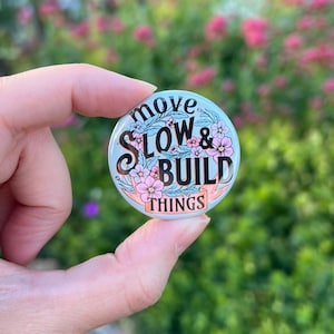 May include: A circular pinback button with a light blue background featuring pink and blue floral illustrations. The text reads move SLOW & BUILD THINGS in black and peach lettering, arranged in a decorative layout with leafy green accents.