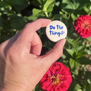 Puede incluir: Una chapa redonda de color blanco con el texto en azul "Do the things!" escrito en una fuente manuscrita informal. La chapa es de forma circular y tiene un acabado liso y mate, mostrando claramente la frase motivadora en su superficie.