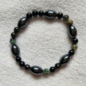 May include: A black and green beaded bracelet with oval-shaped beads. The bracelet is made with black hematite and green aventurine beads.