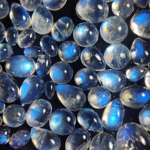 May include: A collection of polished moonstone gemstones in various shapes, including oval, teardrop, and round. The stones display a milky, translucent appearance with blue and white iridescence, set against a dark background.
