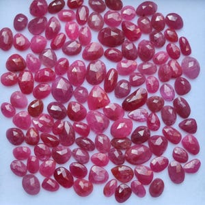 Puede incluir: Una colección de gemas facetadas, de forma ovalada, en tonos rojos y rosas. Las gemas están dispersas sobre una superficie blanca, mostrando sus superficies pulidas y su claridad interna. Las gemas varían en tamaño y forma, algunas más alargadas.