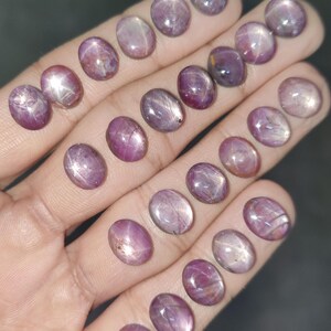Pode incluir: Uma coleção de pedras preciosas de rubi estrela ovais em tons de rosa e roxo. As pedras polidas exibem um efeito de estrela. As gemas são exibidas em uma mão, destacando sua cor e brilho.
