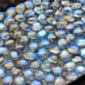 May include: A collection of oval-shaped moonstone cabochons. The stones have a translucent, milky appearance with flashes of blue and white. The polished stones are arranged closely together, creating a shimmering effect. The gemstones are suitable for jewelry making.
