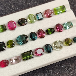May include: Assortment of colorful gemstones in various shapes, including oval, emerald cut, and round. The gems display a range of colors, such as pink, green, blue, and clear, arranged on a white display.