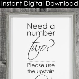 May include: A white sign with black text that reads "Need a number two? Please use the upstairs" with a gray arrow pointing upwards. The sign is framed in a gray frame.