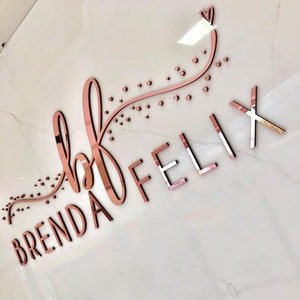 May include: Rose gold metallic sign with the text "Brenda Felix" and a small heart above the "F". The sign has a decorative design of a curved line with small dots.