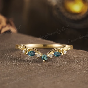 Puede incluir: Un anillo de oro con una banda en forma de V y grabados de hojas. El anillo presenta una piedra preciosa redonda de color verde azulado en el centro, flanqueada por dos piedras de corte marquesa y pequeñas piedras claras a los lados.