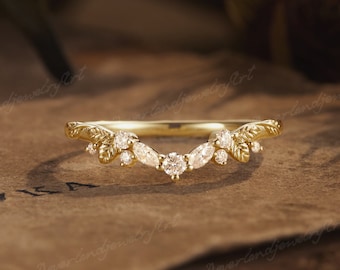 Puede incluir: Un anillo curvo de oro con un diamante redondo central flanqueado por diamantes talla marquesa y redondos, con detalles de hojas grabadas en la banda. El diseño en forma de V incorpora varias piedras claras pequeñas engastadas en metal.