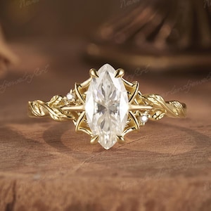 May include: A gold engagement ring with a marquise-cut diamond center stone. The band features a twisted vine design with leaf accents and small accent diamonds. The ring is set against a wooden surface.