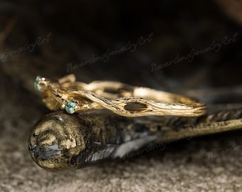 May include: A delicate gold ring with a twig-like design and two small green gemstones. The ring is resting on a textured brown surface.