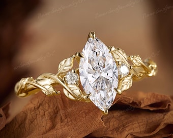 May include: A gold ring featuring a large marquise-cut clear gemstone at the centre, flanked by two smaller round-cut clear stones. The band is designed with a detailed vine and leaf motif that wraps around the finger in a continuous pattern.
