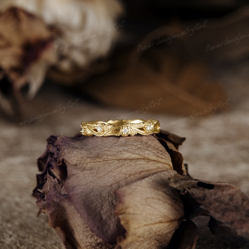 Puede incluir: Una delicada banda de oro con un dise&ntilde;o floral y peque&ntilde;os diamantes. El anillo est&aacute; colocado sobre una rosa seca.