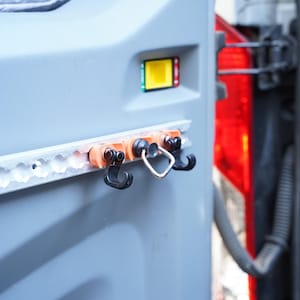 Könnte beinhalten: Nahaufnahme eines silbernen Metallschienen mit drei schwarzen Haken und einem silbernen Ring, der daran befestigt ist. Die Schiene ist an einer grauen Oberfläche montiert.