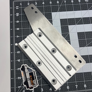 May include: Two silver metal pieces with multiple holes, one rectangular and one elongated, are positioned on a black and gray grid surface. A sticker with a van graphic and the text "GET KNARLY" is also present. The items appear to be mechanical components.