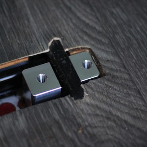May include: Close-up of two silver metal brackets with threaded holes, secured with black tape within a recessed area of a dark wood-grain surface. The brackets are square and appear to be for mounting or securing something.