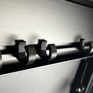 May include: Five black plastic hooks mounted on a silver metal bar. The hooks are attached to a black surface.