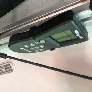 May include: Black MAX-AIR remote control with a small LCD screen and several buttons, including "FAN ON/OFF." The remote is mounted on a metal rail with a black strap. The background is a light-colored interior.